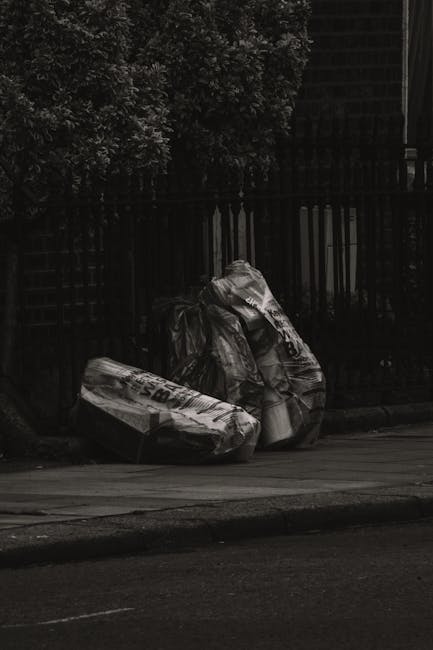 A man with dark hair and wearing a black T-shirt with white graphic text is seen disposing of rubbish into a dedicated metal waste bin on a paved sidewalk. The bin is cylindrical, made of stainless st