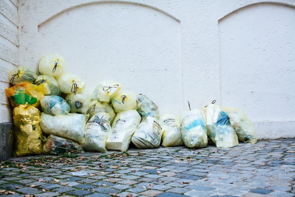 A middle-aged man with dark hair wearing a black t-shirt and dark trousers is seen disposing of trash into a metallic recycling bin on a paved walkway. He is holding a large white waste bag filled wit